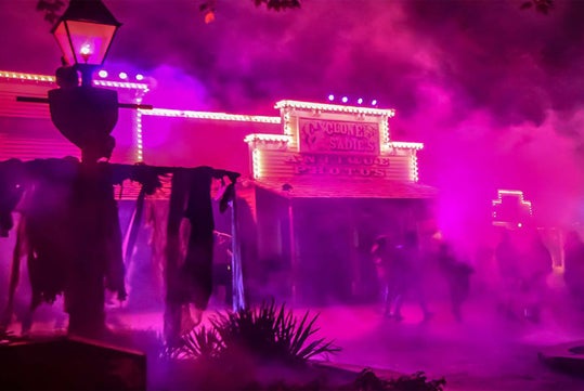 Foggy scene at night with neon-lit buildings and people walking, featuring old western-style architecture and hanging fabric decorations.