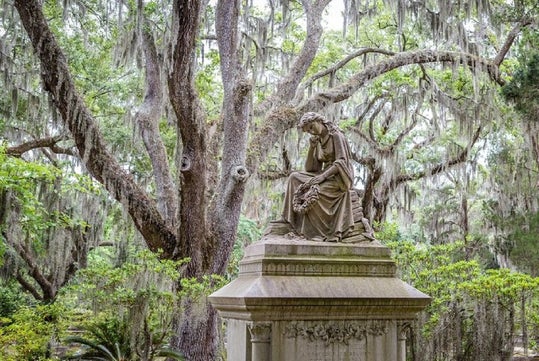 A stone statue of a seated figure atop a pedestal, surrounded by lush greenery and large trees draped with Spanish moss.