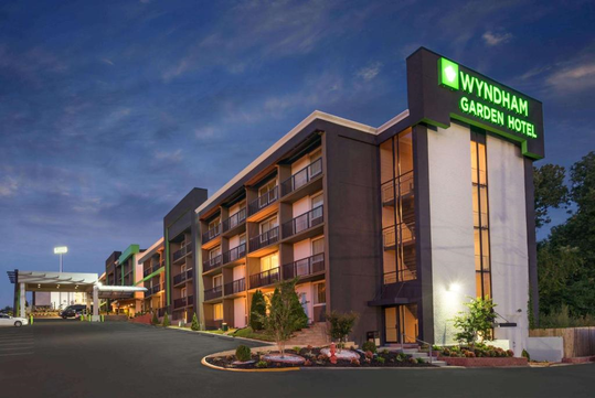 Exterior view of the Wyndham Garden Hotel at dusk, with a landscaped entrance and a lit signage on the building.