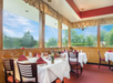 A restaurant dining area with several tables set with white tablecloths, wine glasses, and folded red napkins. Large windows provide a view of green trees outside.