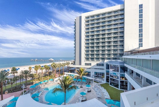A tall beachfront hotel with balconies overlooks a pool area surrounded by palm trees, with the ocean and a pier visible in the background under a blue sky with wispy clouds.