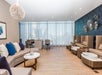 Modern spa lounge with beige seating, blue accent wall, manicure tables, and pedicure chairs, featuring large windows with sheer curtains and wooden floors.