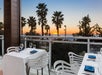 Outdoor restaurant tables with white tablecloths and glasses overlook palm trees and the ocean at sunset, seen through a glass railing.