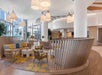 Modern hotel lobby with curved seating, round tables, armchairs, large white columns, pendant lights, and floor-to-ceiling windows letting in natural light.