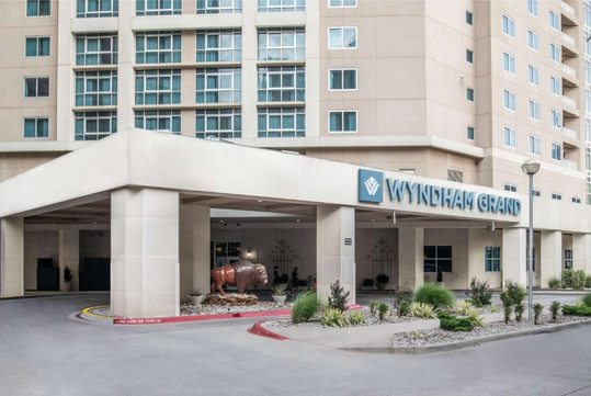 Entrance of a hotel with "Wyndham Grand" sign, featuring a revolving door, landscaping, and a sculpture of a bison in front.
