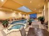 Indoor hotel pool area with a hot tub, lounge chairs, tables, and large potted plants. The ceiling has a skylight. The floor is tiled, and the room is well-lit.