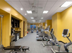 A gym room with yellow walls features treadmills, exercise bikes, a weight bench, and a cable machine. A refrigerator and a shelf with towels are on the left.