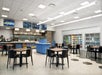 A modern cafeteria with black tables and chairs, a counter with menu boards above, a visible kitchen, and refrigerated drink coolers along the wall.