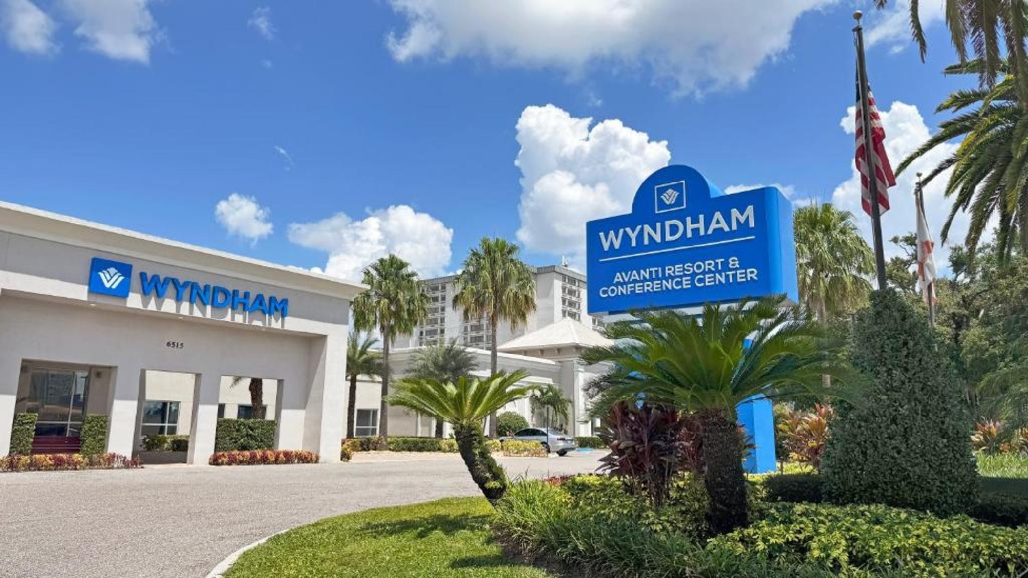 Entrance of the Wyndham Avanti Resort & Conference Center with a blue sign, palm trees, and a driveway under a sunny sky.