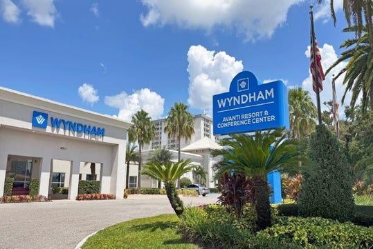 Entrance of the Wyndham Avanti Resort & Conference Center with a blue sign, palm trees, and a driveway under a sunny sky.