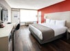 Hotel room with a king bed, red accent wall, desk with chair, flat-screen TV, and large window letting in natural light.