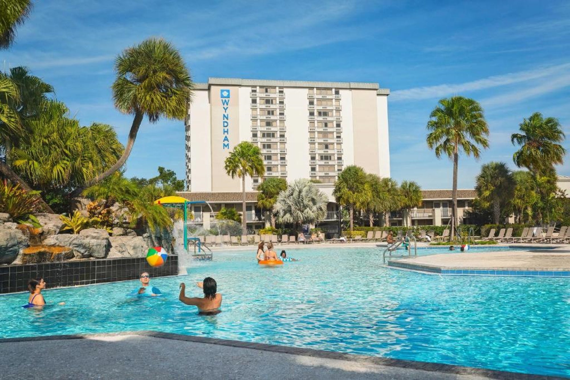 People swim and play in a large outdoor pool surrounded by palm trees, with a Wyndham hotel building visible in the background.