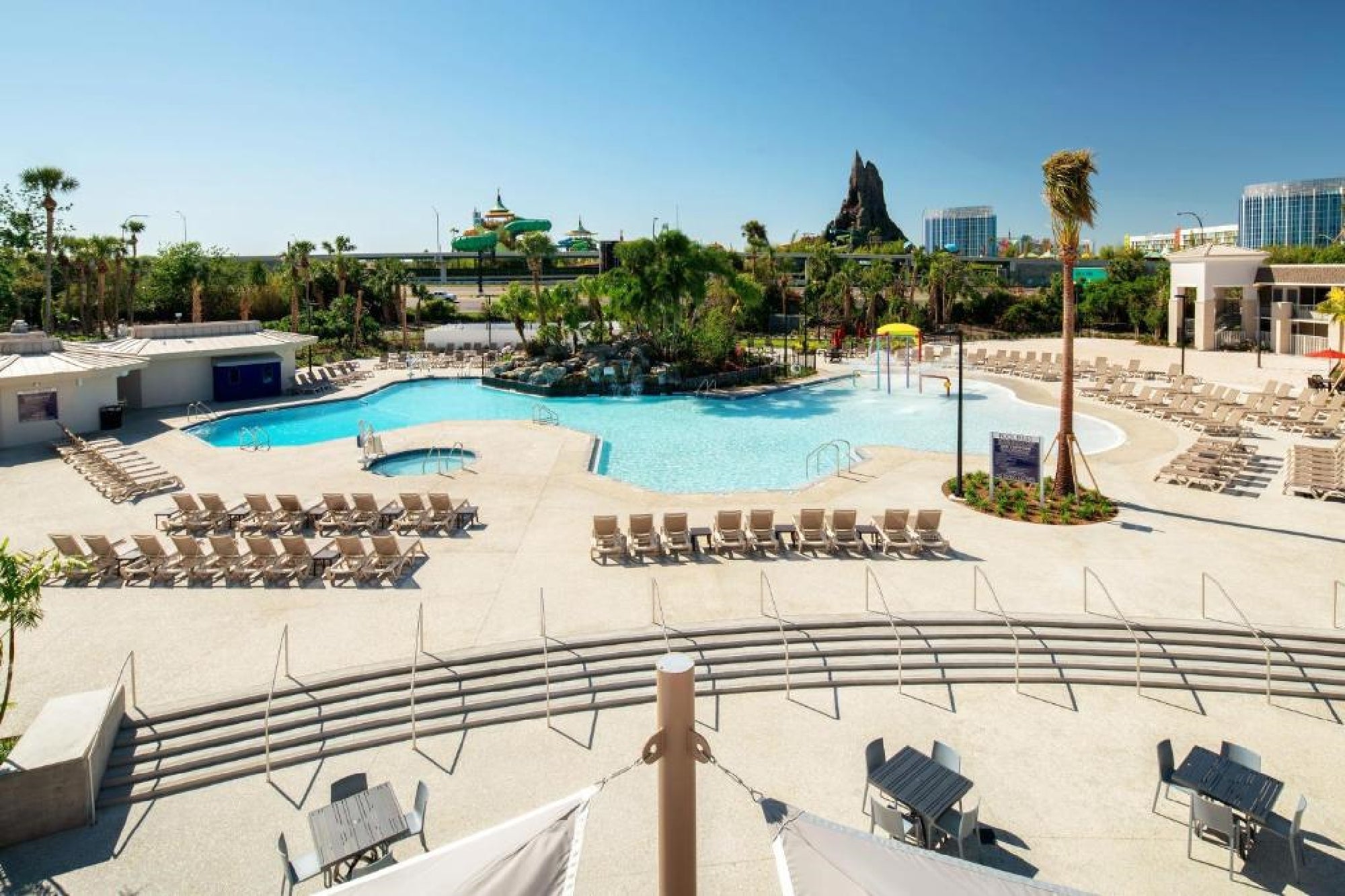 Large outdoor swimming pool area with lounge chairs, palm trees, and water features, set against a backdrop of resort buildings and a distant volcano-shaped structure.