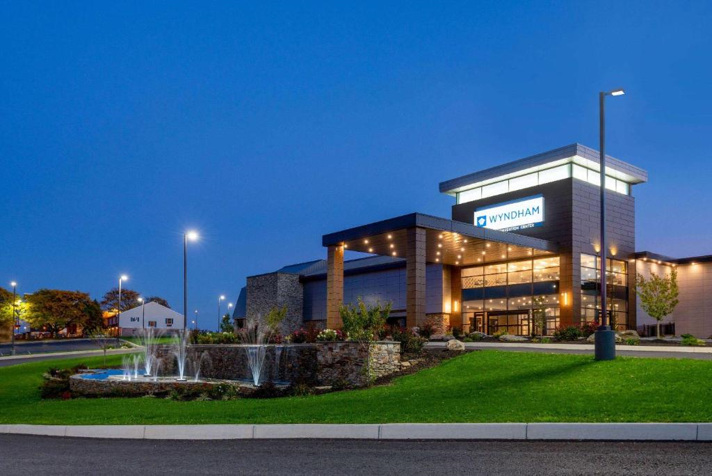 Modern hotel building with stone pillars and large windows, sign reading "Wyndham." A fountain and landscaped garden are in the foreground. It's evening with a clear sky.