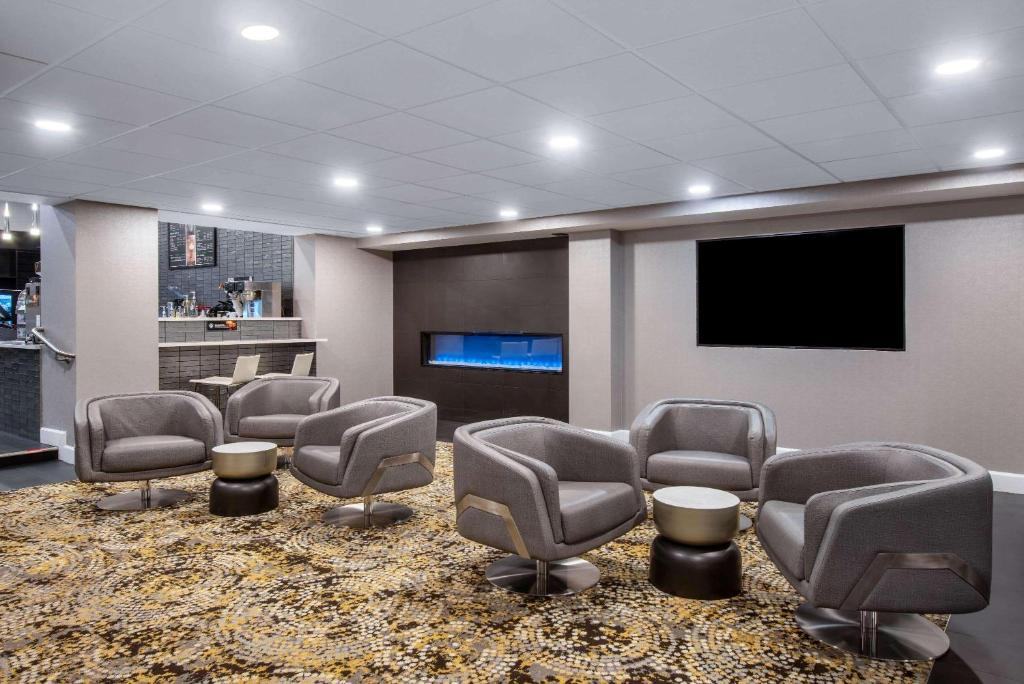 Modern lounge area with six gray armchairs, round side tables, a wall-mounted TV, and a built-in fireplace. The ceiling has recessed lighting, and the carpet features a patterned design.