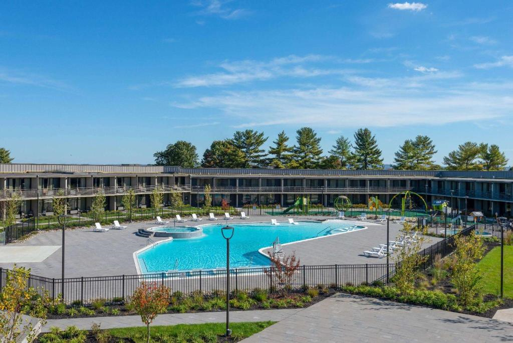 Outdoor view of a hotel courtyard featuring a large, oval-shaped swimming pool surrounded by lounge chairs within a landscaped area. A two-story building encircles the pool.