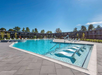 Rectangular outdoor swimming pool with submerged loungers, surrounded by a paved deck and a multi-story building in the background, under a clear blue sky.
