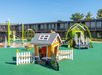 A colorful playground with playhouses, slides, and climbing structures on rubber flooring, surrounded by a two-story building under a clear blue sky.