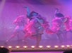 Three dancers perform on stage under pink lights, wearing pink costumes and fishnet stockings, with a sparkly backdrop.
