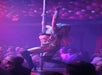 Performer in a leopard print outfit and boots dances on a pole on stage, with an audience seated in the background.