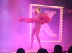 Performer on stage in a pink dress and feather boa, surrounded by pink lights and a lit-up letter "X" in the background.