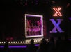 Performers on stage lined up behind a lit rectangle, with a large illuminated "X" beside them, in front of an audience.