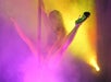 A person performs a pole dance move, balancing with legs extended, surrounded by colorful stage lights and mist.