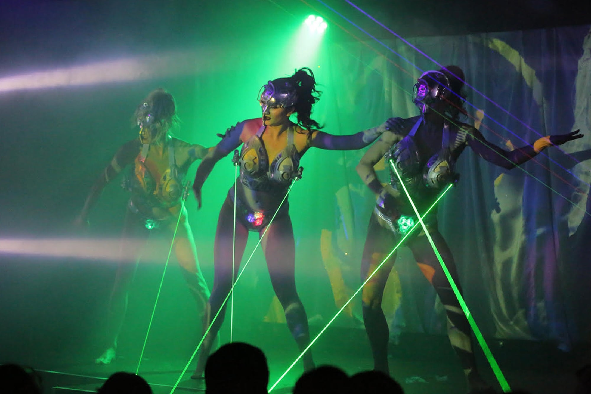 Three performers in body paint and masks pose under green stage lights and lasers during a theatrical show.