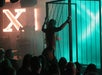 A performer dances inside a metal cage on stage, illuminated by teal and orange lights, while an audience watches in a dimly lit venue.