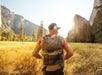 A person wearing a backpack stands in a grassy valley surrounded by tall trees and mountains with the sun shining in the background.