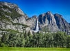 Large waterfall cascades down a rocky mountainside into a forested area under a clear blue sky, with a grassy field in the foreground.