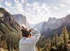 A person in a knit hat and jacket photographs a mountainous valley covered with trees under a partly cloudy sky.