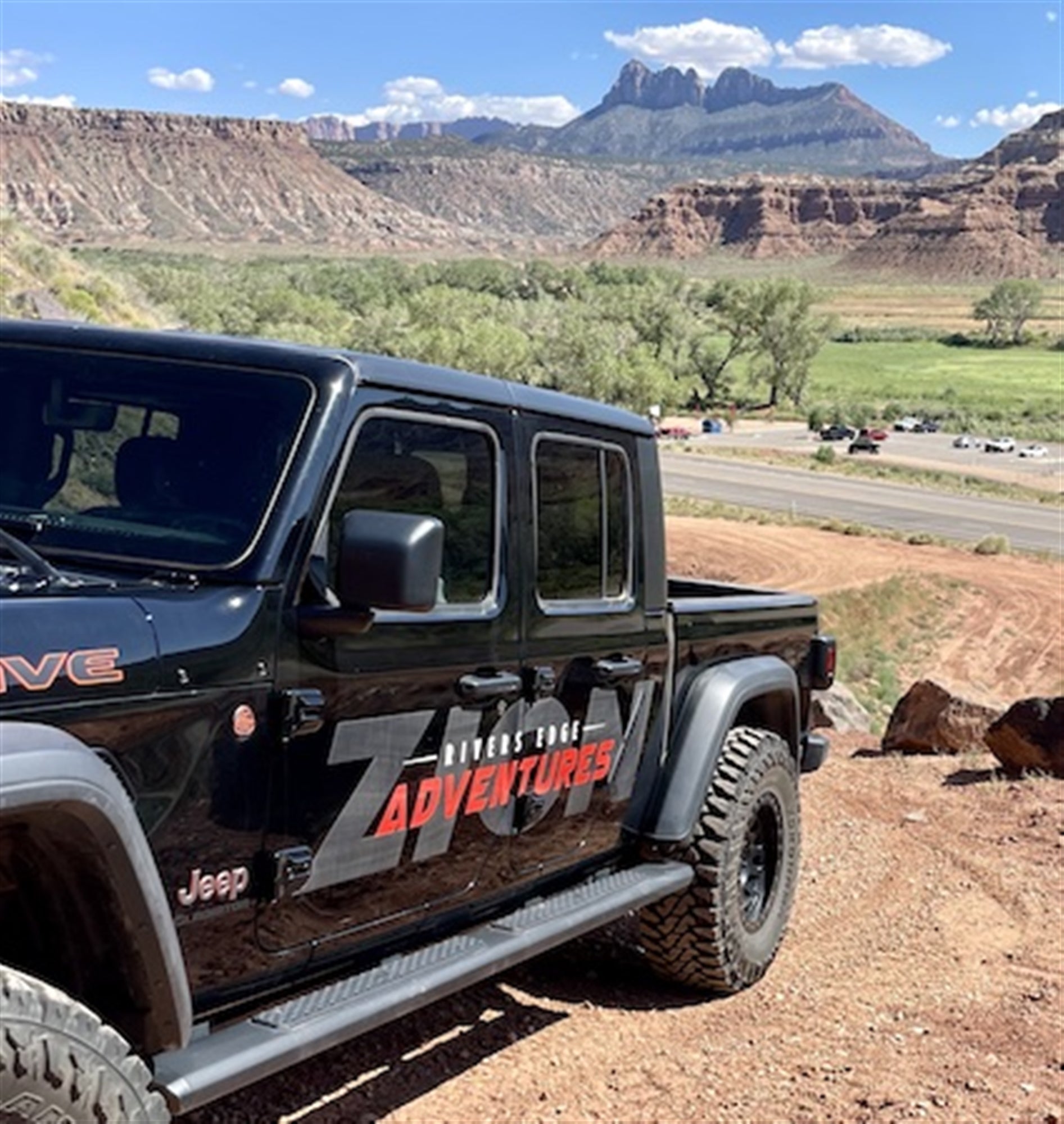 A black Jeep Gladiator with "Zion Adventures" branding is parked on a red dirt road with rocky cliffs and mountains in the background on a sunny day.