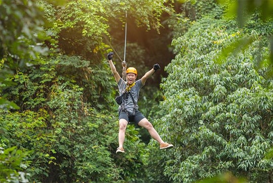 Person wearing a helmet and harness zip-lining through a green forest, smiling and raising one arm in excitement.