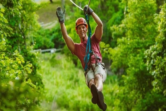 Person zip-lining through a lush green forest, wearing a helmet, gloves, and harness, with one arm raised.