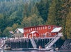 A red and green building sits on a wooden pier with boats docked in front, surrounded by dense forest and tall trees.