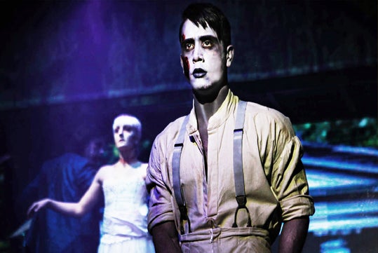 A person with white face paint and dark makeup stands in the foreground, wearing suspenders and a light shirt; another person in a white dress stands blurred in the background.