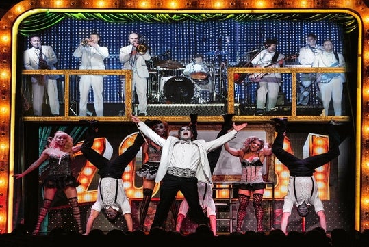 A man in a white suit and zombie makeup standing center stage with men doing handstands and women in lingerie behind him.