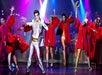 A man in a metallic suit sings and dances on stage with several performers in red costumes under colorful stage lights.