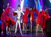 A man in a white suit and zombie makeup with women and men in red robes and zombie makeup behind him.