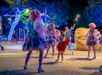 Performers in colorful costumes and wigs dance around a child in a red dress at an outdoor area decorated with illuminated trees at night.