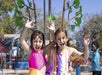 Two young girls in swimsuits smile and raise their hands while playing at a water park with sprays and colorful equipment in the background.