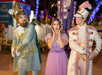 Three people in elaborate costumes pose outdoors at night; one wears a blue and gold suit, one a purple dress, and one a white and pink uniform with feathered hat.