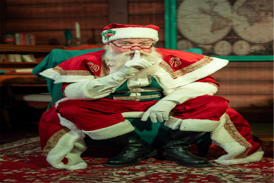 A person dressed as Santa Claus sits on a chair in a festive room, holding a finger to their lips in a shushing gesture.