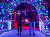 Two adults and a child walk under a tunnel of colorful holiday lights at night. The adults swing the child by the arms, and all are wearing red plaid shirts.