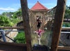 A young child crosses a rope bridge toward the camera, followed by two adults, in an outdoor playground surrounded by greenery.