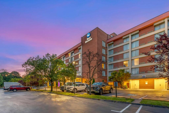 Hotel exterior with a Best Western Plus sign, five-story brick building, parked cars, and trees in the foreground at sunset.