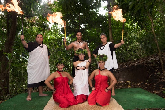 A group of people in traditional attire, with three holding fire torches and two wearing red dresses, pose outdoors surrounded by greenery.