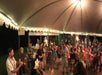 A group of people stand under a tent, participating in a dance or activity. The scene is lit by string lights and a chandelier.