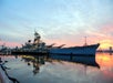 A large ship in the water at the Battleship New Jersey Museum and Memorial, seen against a backdrop of the sky and water, with reflections in the water. The scene captures the ship during either sunrise or sunset.
